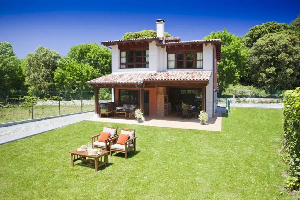 Casa rural de dos plantas, con amplio jardín verde con una mesita y dos sillones