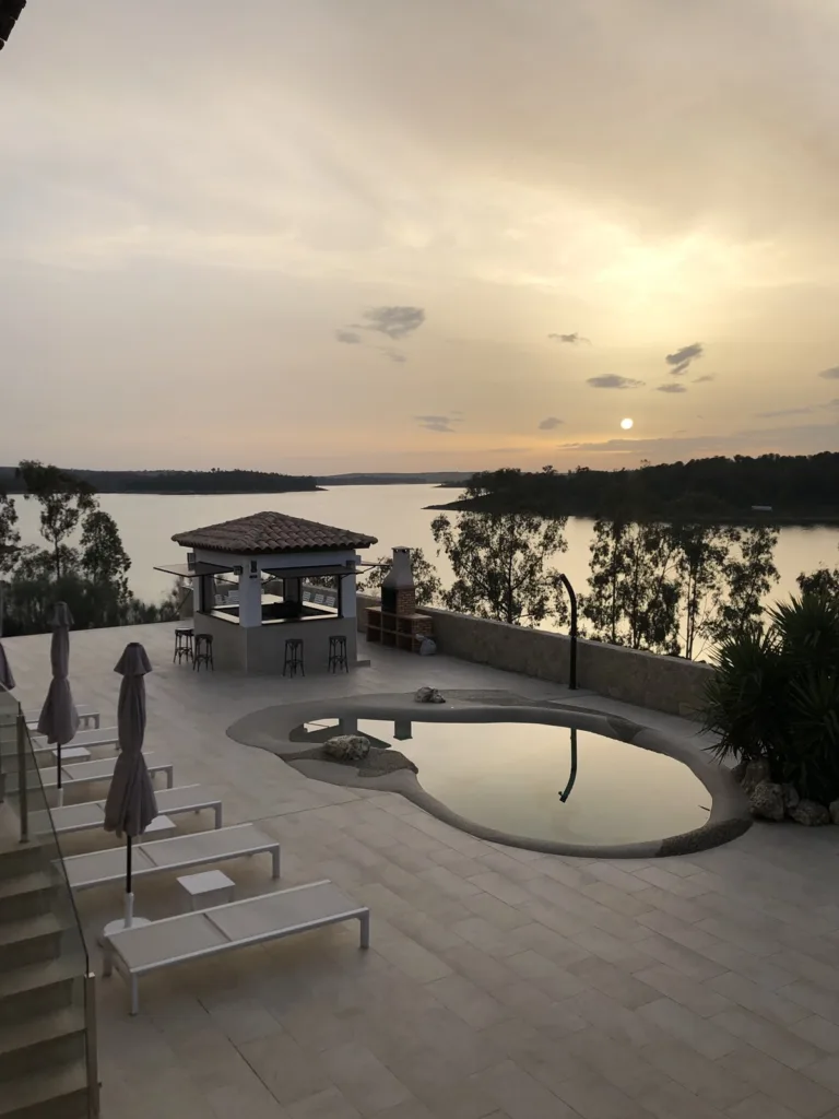 Puesta del sol desde una terraza con hamacas, piscina y un embalse al fondo