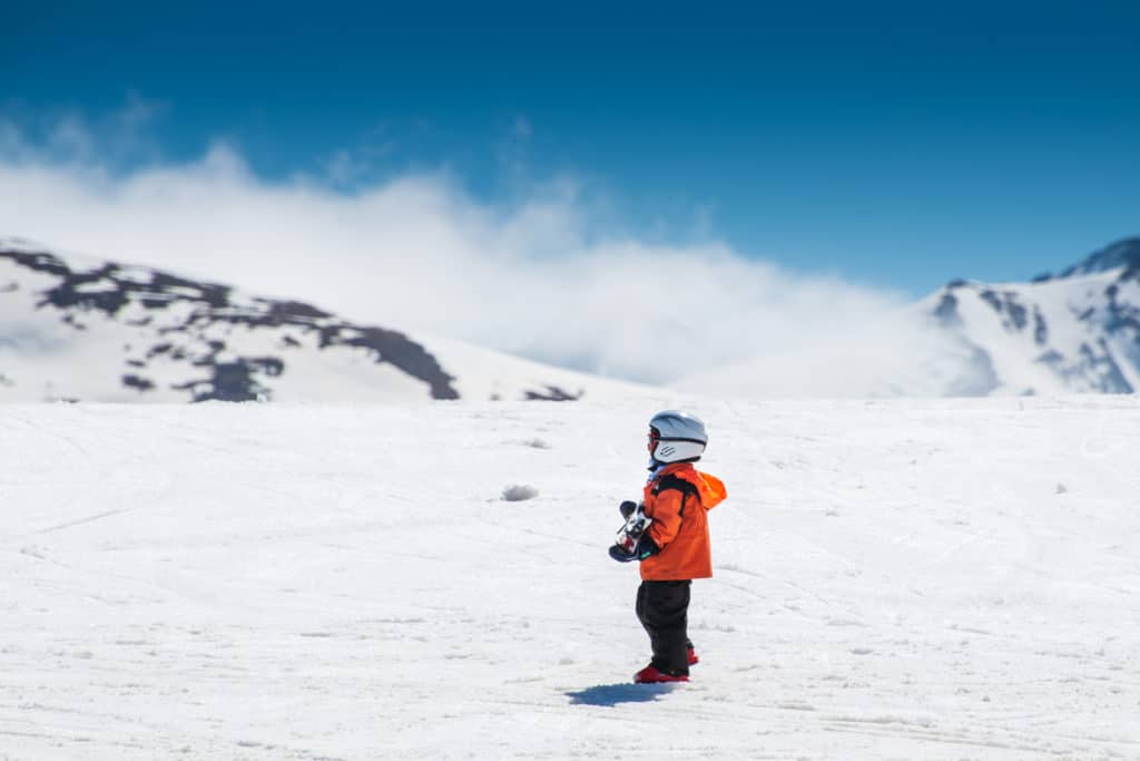 Un niño pequeño con casco, chaqueta naranja y pantalones oscuros está de pie en una ladera nevada, sujetando unos bastones de esquí, con montañas nevadas y un cielo azul de fondo.