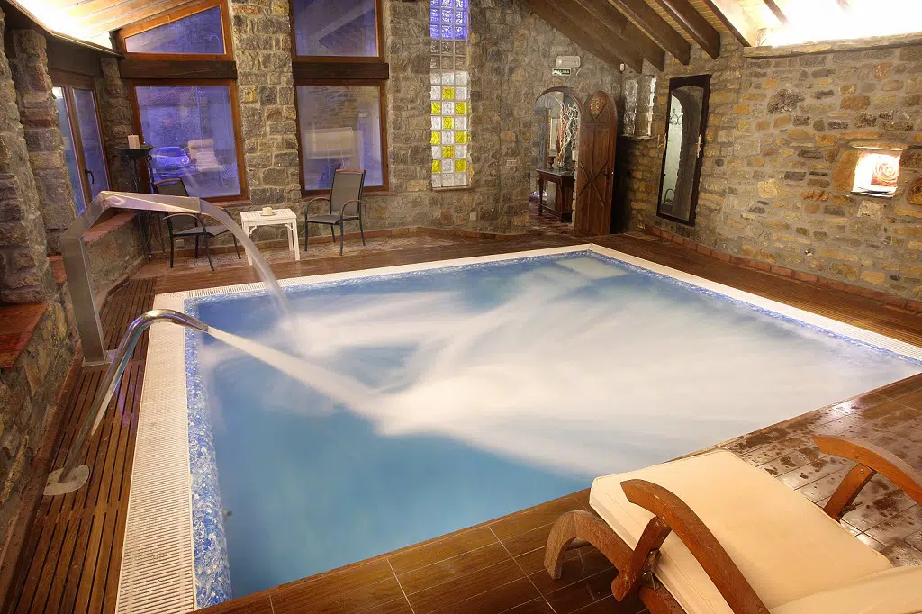 Acogedora piscina cubierta en una sala rústica con paredes de piedra, con chorros de agua, tumbonas de madera y una cálida iluminación: una lujosa casa rural con piscina climatizada para la máxima relajación.