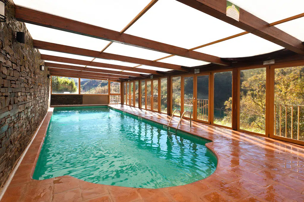 Esta casa rural con piscina climatizada cuenta con una piscina cubierta de agua cristalina, rodeada por un suelo de baldosas rojas y un muro de piedra, con grandes ventanales de cristal que revelan impresionantes vistas de los árboles y las colinas del exterior.