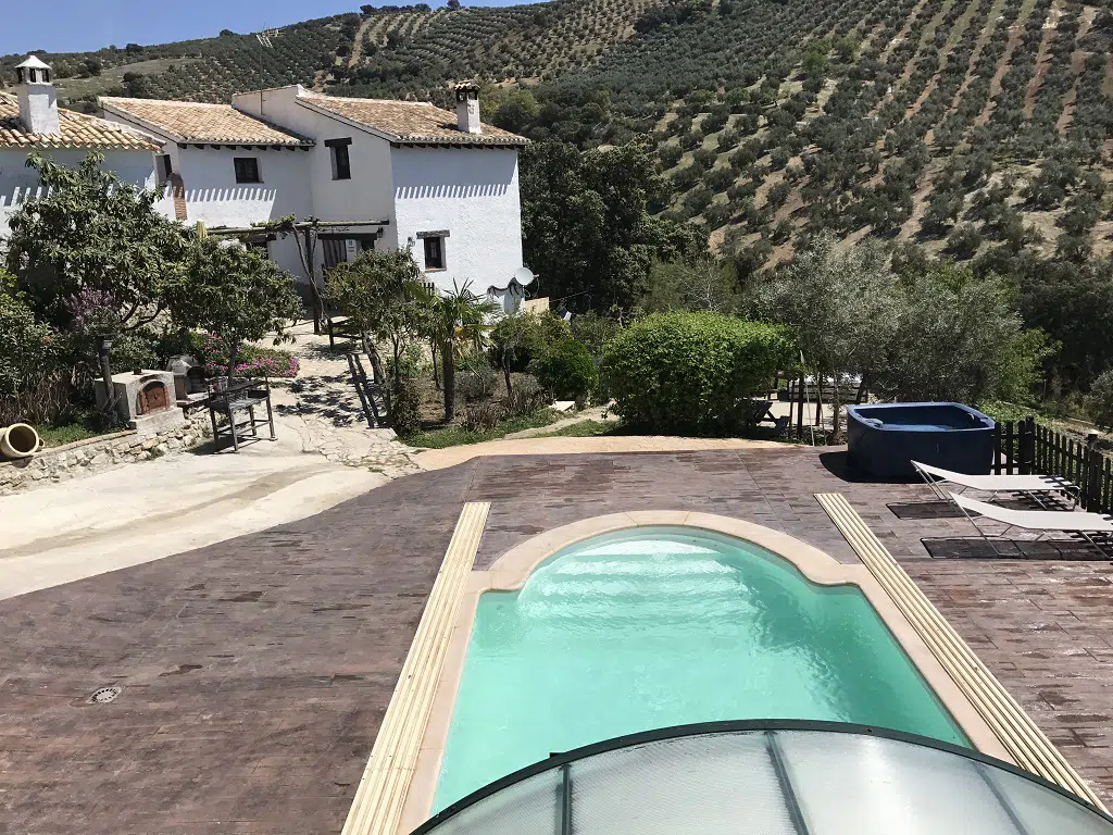Una casa rural con piscina climatizada y bañera de hidromasaje se encuentra en un patio junto a una casa blanca con tejado de tejas, rodeada de árboles y arbustos verdes, con vistas a colinas en terrazas y olivares al fondo.