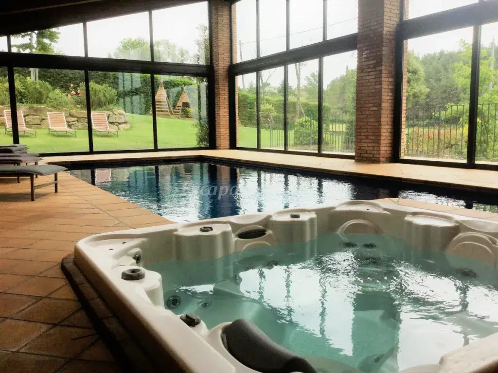 Zona de piscina cubierta con grandes ventanales con vistas a un verde jardín; en primer plano hay una bañera de hidromasaje y en el exterior tumbonas. Disfrute de la luz natural en esta espaciosa y moderna casa rural con piscina climatizada.