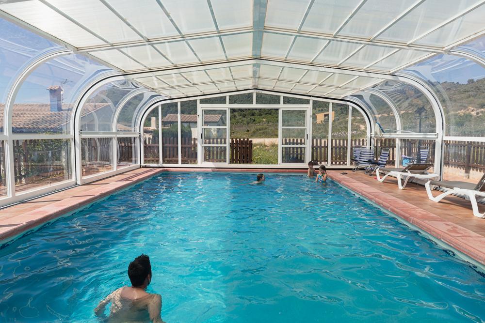 Una piscina cubierta con techo transparente y curvado aporta a esta casa rural con piscina climatizada un ambiente relajante. Tres personas disfrutan del agua caliente mientras la luz del sol entra a raudales, mientras las tumbonas vacías y las vistas a la exuberante vegetación completan la escena.