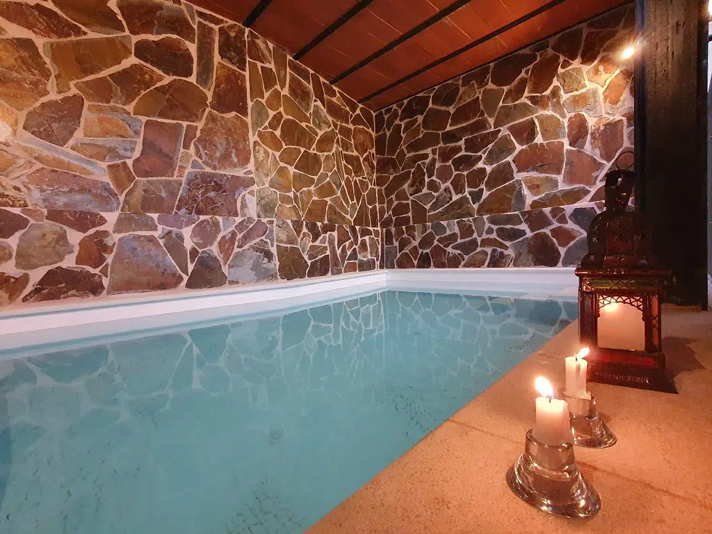 Una acogedora casa rural con piscina climatizada cuenta con paredes de piedra, techo de madera e iluminación tenue. Tres velas encendidas y un farol decorativo junto a la piscina crean un ambiente relajante y tranquilo.