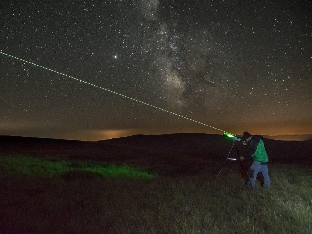 Una persona utiliza un puntero láser verde apuntando al cielo nocturno lleno de estrellas, con la Vía Láctea visible por encima de colinas oscuras y suelo cubierto de hierba, bajo un cielo claro y estrellado.