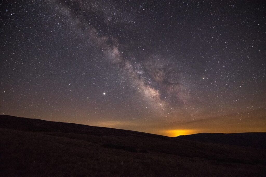 Un cielo nocturno lleno de estrellas con la Vía Láctea brilla sobre un paisaje oscuro y montañoso, perfecto para una escapada rural. En el horizonte aparece un tenue resplandor anaranjado que contrasta a la perfección con el tranquilo primer plano.