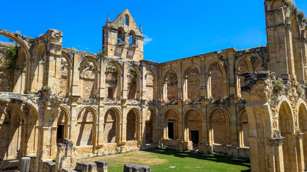 Antiguas ruinas de piedra de un gran edificio con ventanas arqueadas, columnas y un campanario parcialmente en pie bajo un cielo azul brillante, con hierba verde cubriendo el suelo en su interior.