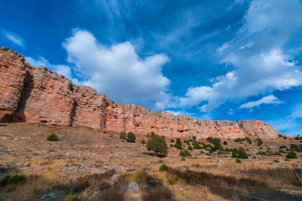 Un amplio acantilado rocoso se eleva sobre un paisaje seco y herboso salpicado de pequeños arbustos, perfecto para el turismo rural, bajo un cielo azul brillante repleto de grandes nubes blancas.