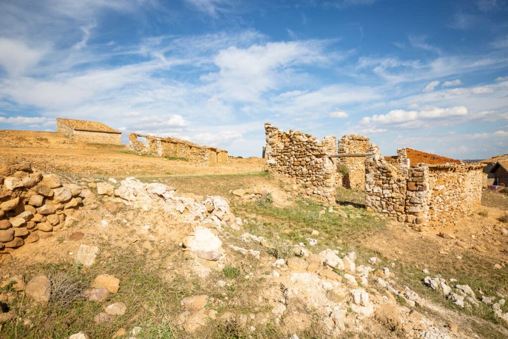 Ruinas de antiguos edificios de piedra en una ladera seca y cubierta de hierba bajo un cielo azul con nubes dispersas. Las estructuras están parcialmente derruidas, rodeadas de rocas y parches de vegetación.