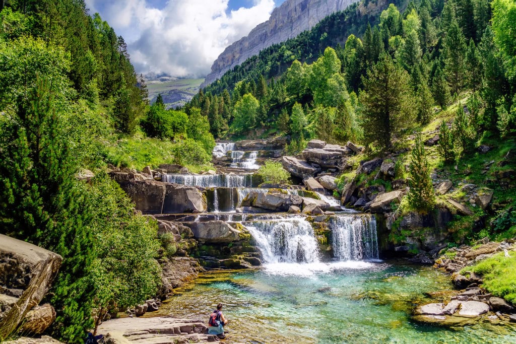 Un excursionista se sienta junto a una poza de aguas claras y turquesas en la base de la Cascada Cola de Caballo, rodeado de frondosos árboles verdes y acantilados rocosos bajo un cielo parcialmente nublado en un impresionante paisaje montañoso.