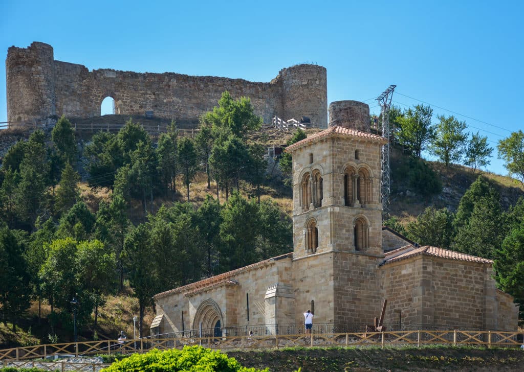 Una iglesia de piedra con un campanario se alza ante una colina boscosa, invitando a los visitantes a disfrutar del turismo rural, mientras las ruinas de una antigua fortaleza coronan la cima de la colina bajo un cielo azul despejado.