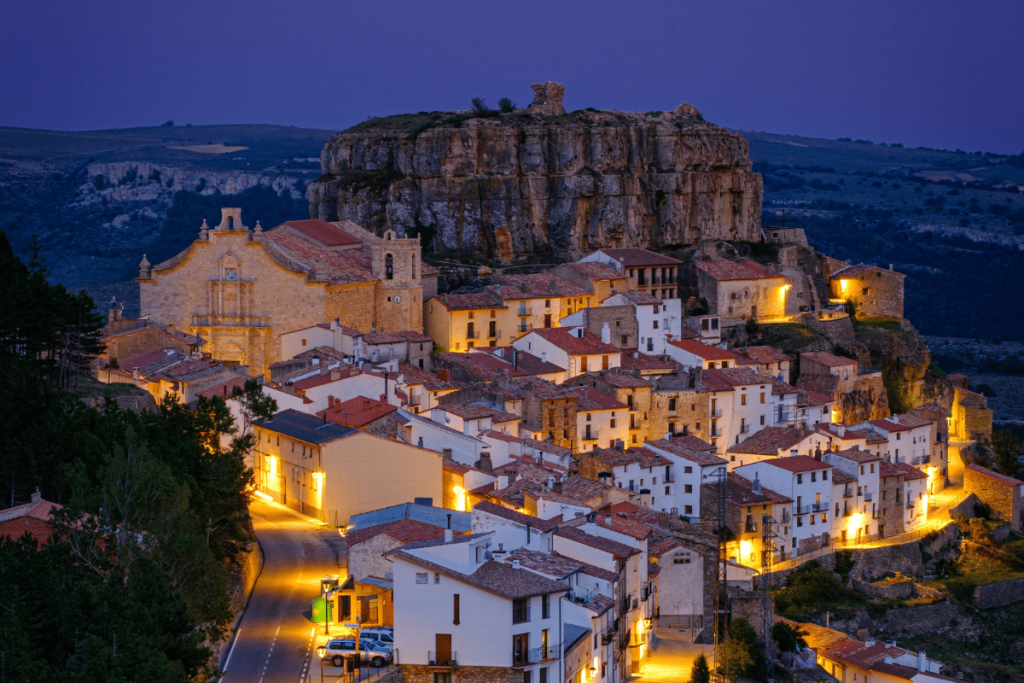 Pueblo en lo alto de una colina al anochecer, con cálidas farolas que iluminan casas de tejados blancos y rojos, una gran iglesia y un acantilado rocoso coronado por antiguas ruinas al fondo. A lo lejos se ven colinas onduladas.