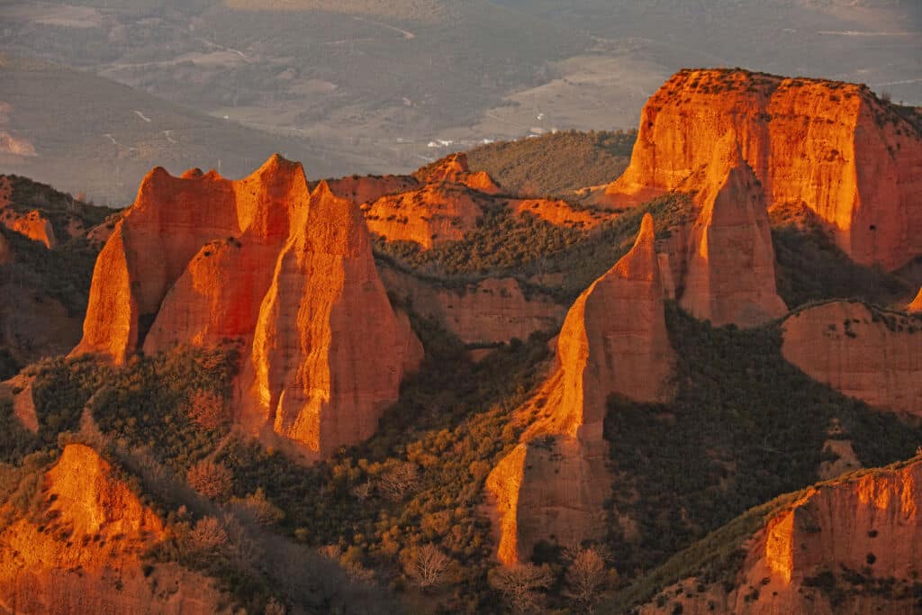 Llamativas formaciones rocosas de color rojo anaranjado con picos escarpados y dentados se elevan sobre la verde vegetación bajo la cálida luz del sol, con un telón de fondo de colinas onduladas y campos lejanos.