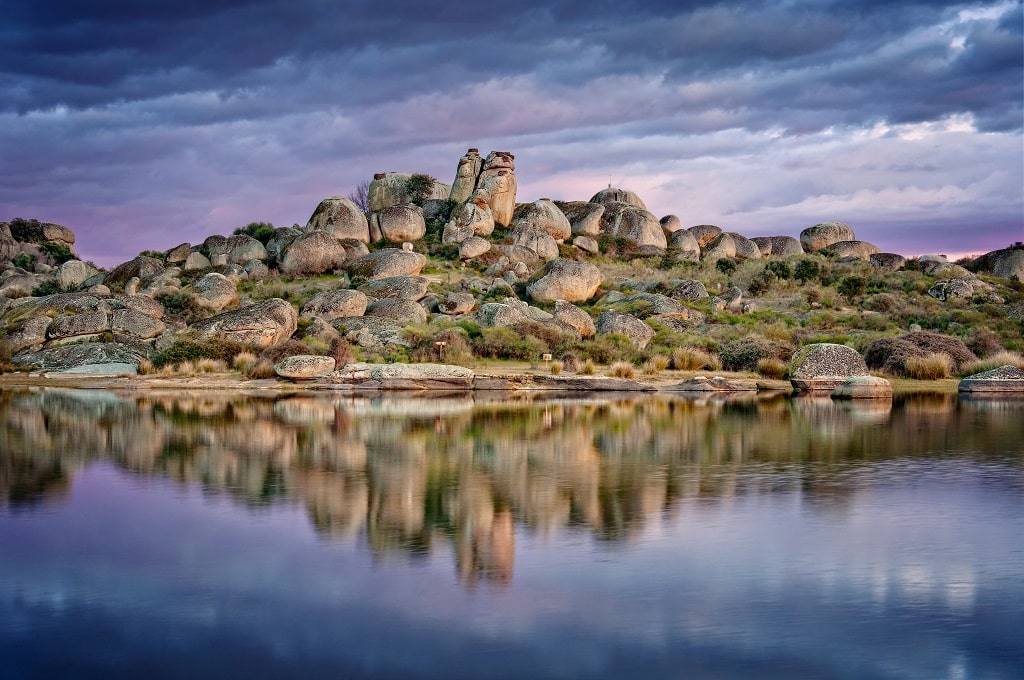 Grandes rocas redondeadas se asientan sobre una colina rocosa, reflejadas en las tranquilas y quietas aguas de abajo. El cielo está nublado con toques de púrpura y azul, creando un paisaje sereno y tranquilo.