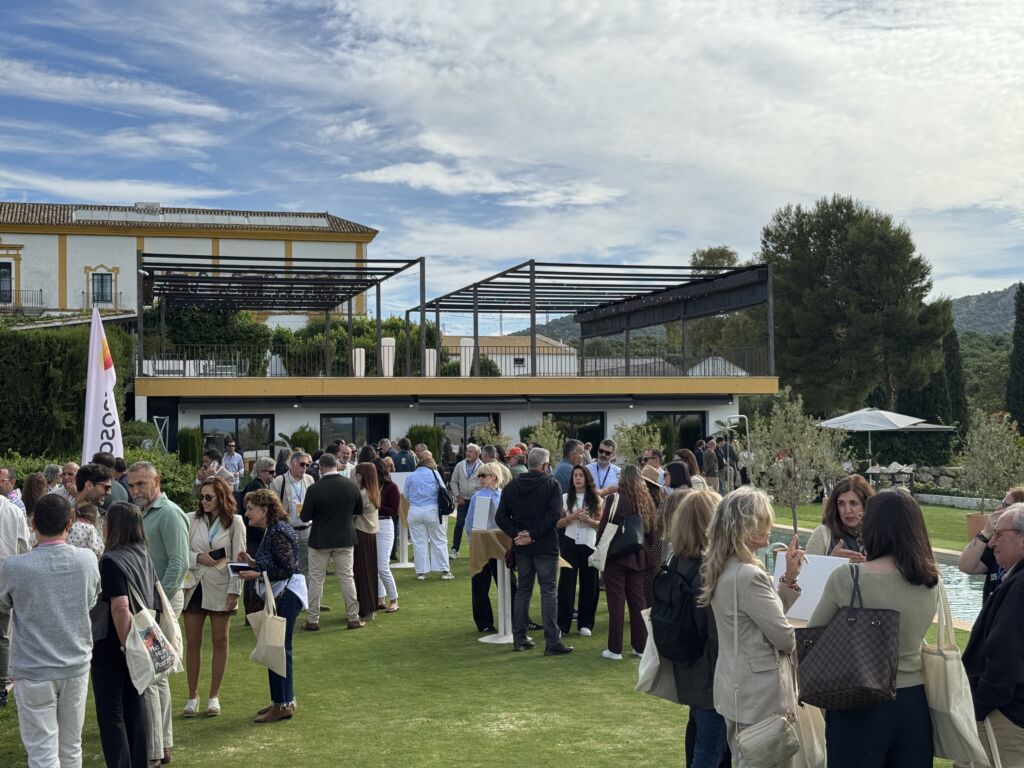 Un grupo numeroso de personas socializa y establece contactos al aire libre en un prado cubierto de hierba frente a unos edificios modernos, bajo un cielo parcialmente nublado. Muchos llevan bolsas de mano y visten ropa informal de negocios. La zona está rodeada de árboles y jardines.