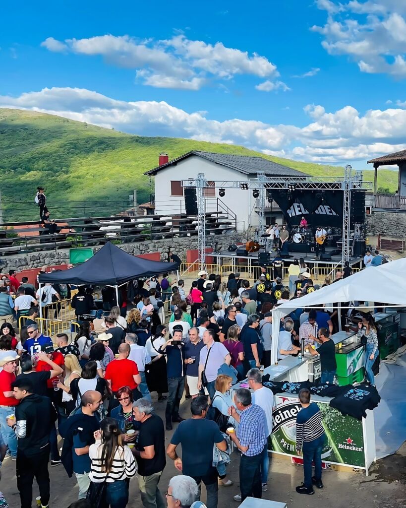 Un animado festival de música al aire libre con una multitud frente a un escenario, puestos de comida y bebida, montañas al fondo y un cielo parcialmente nublado. La gente socializa y disfruta del evento.