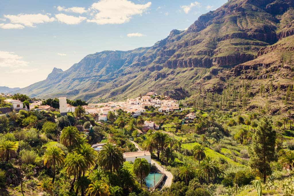Un pequeño pueblo de edificios blancos y tejados naranjas está enclavado en un exuberante valle verde rodeado de altas y escarpadas montañas bajo un cielo casi despejado. El paisaje está lleno de palmeras y vegetación.