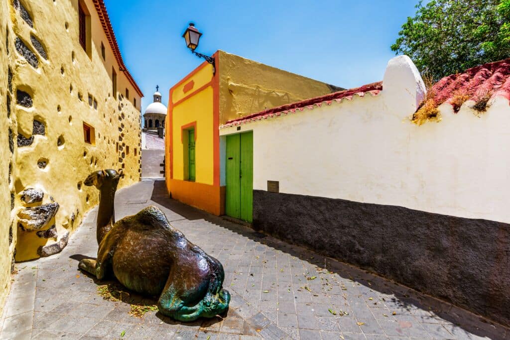 Una calle estrecha y soleada bordeada de edificios amarillos y blancos presenta una estatua de bronce de un camello tumbado sobre los adoquines. Una puerta verde y una cúpula blanca se ven al fondo bajo un cielo azul despejado.