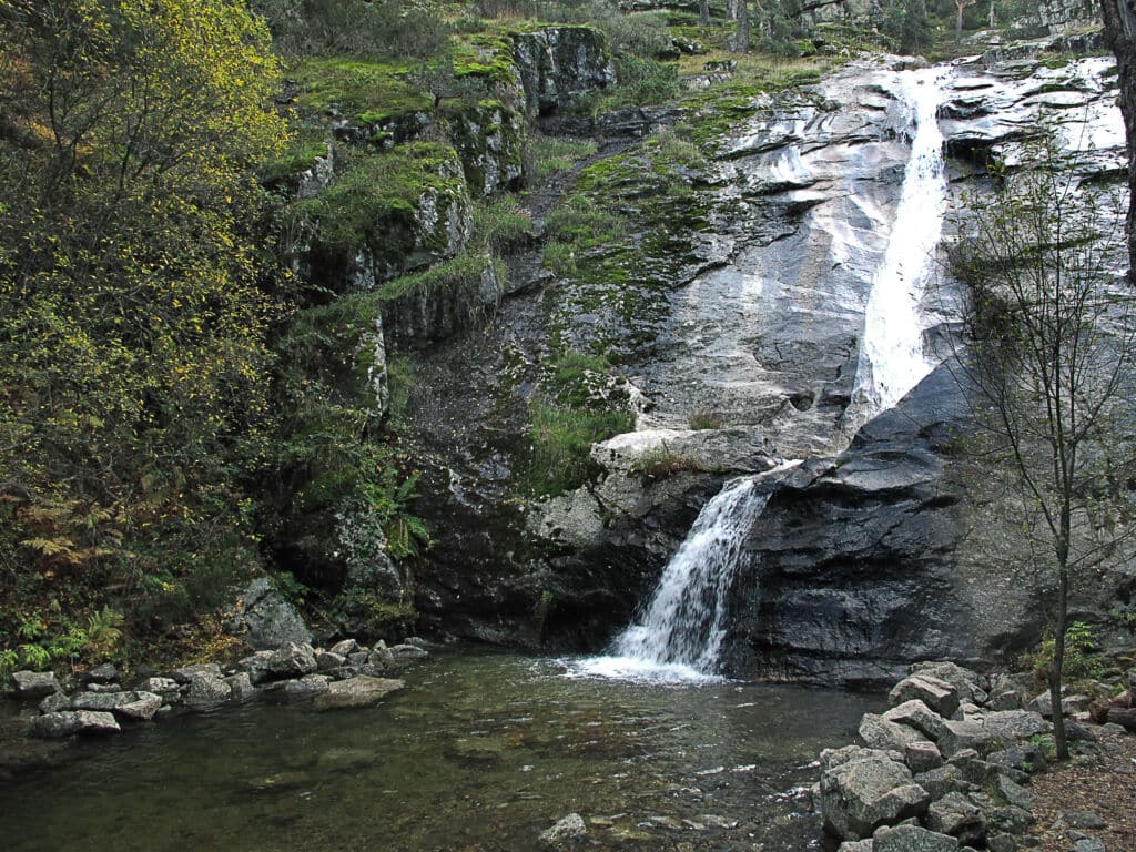 Una pequeña cascada cae sobre rocas musgosas en un estanque transparente, rodeado de verdes árboles y escarpados acantilados de piedra en una zona boscosa.
