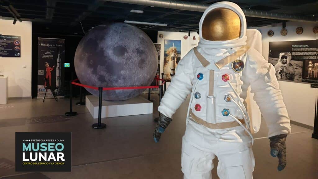 Una figura de un astronauta a tamaño real se encuentra frente a una gran maqueta de la Luna y a varios paneles informativos en el Museo Lunar, un museo dedicado al espacio y a la ciencia. La zona está acordonada y el logotipo del museo aparece en la esquina inferior izquierda.
