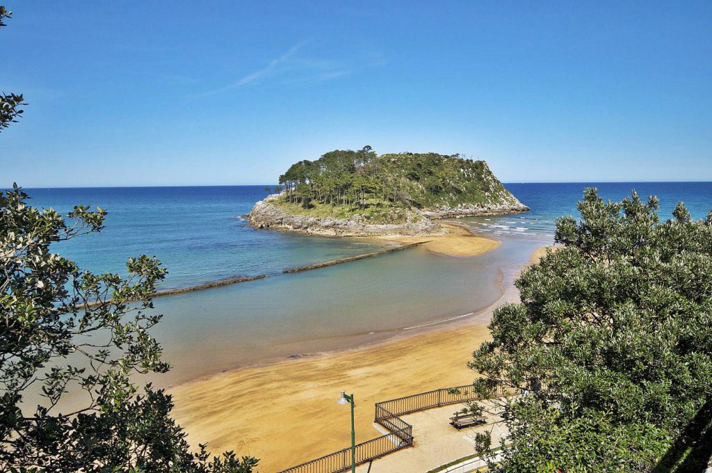 Una pequeña isla cubierta de árboles se asienta cerca de la orilla arenosa, separada por un estrecho banco de arena en aguas azules y transparentes. El verde follaje enmarca la escena, y una valla de madera bordea un sendero a lo largo de la costa.